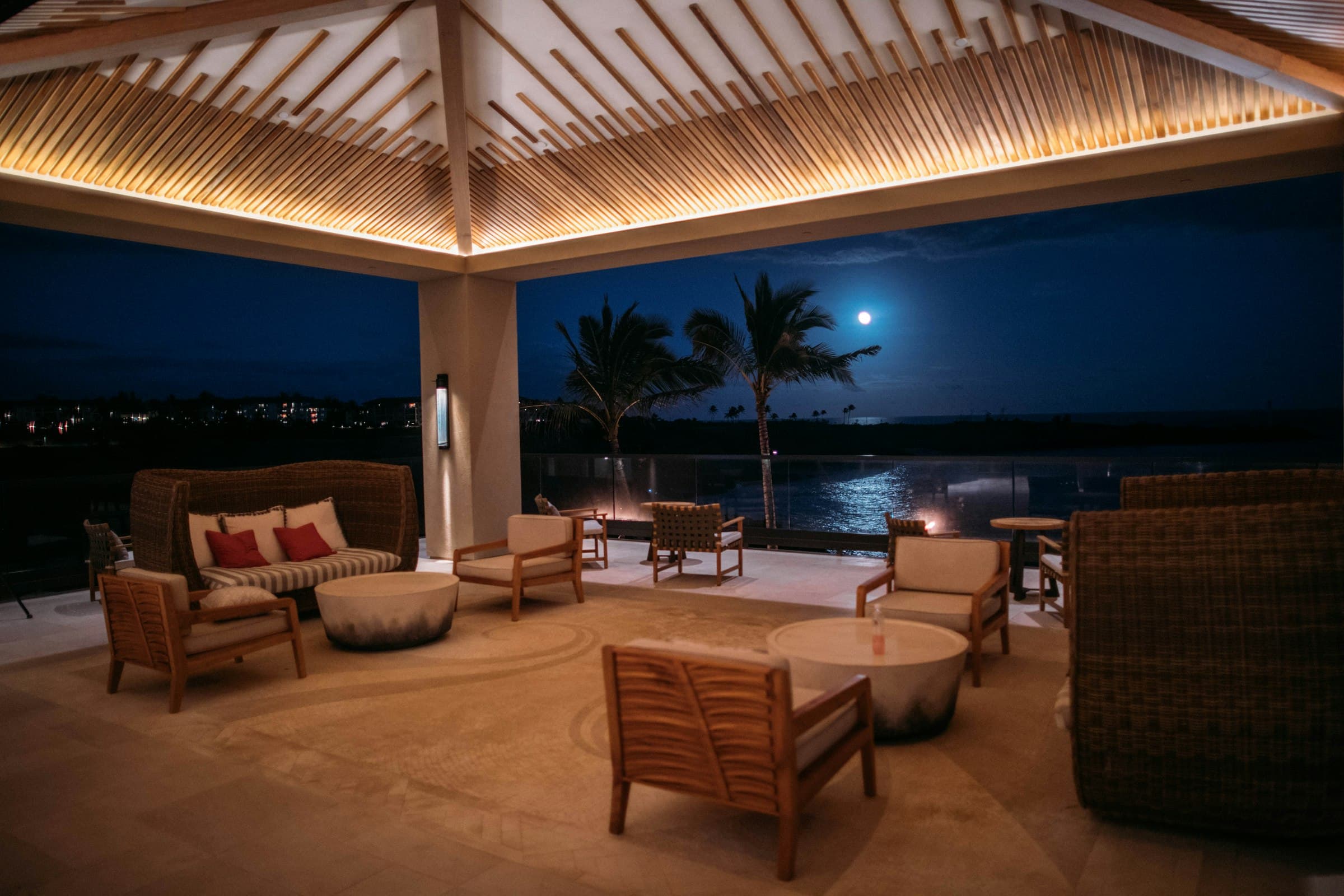 An open-air pavilion at night with a cove-lit slatted ceiling and the moon over the water